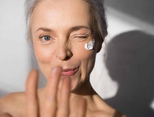 Portrait view of the friendly mature woman with protective skin care cream and moisturizer lotion on her happy smiling face posing at the studio and showing her hand to the camera